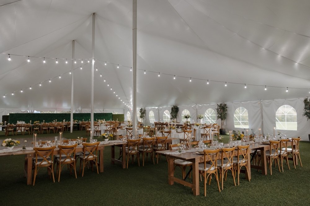 Elegant wedding reception setup inside a large white tent with string lights and wooden tables and chairs
